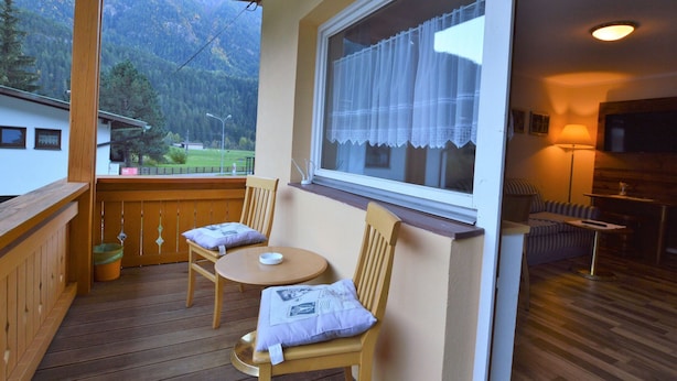 A wooden deck with two chairs and a small table, a window with a curtain, and a view of mountains. - Apartment in Oetz Near Ski Lift (Oetz)