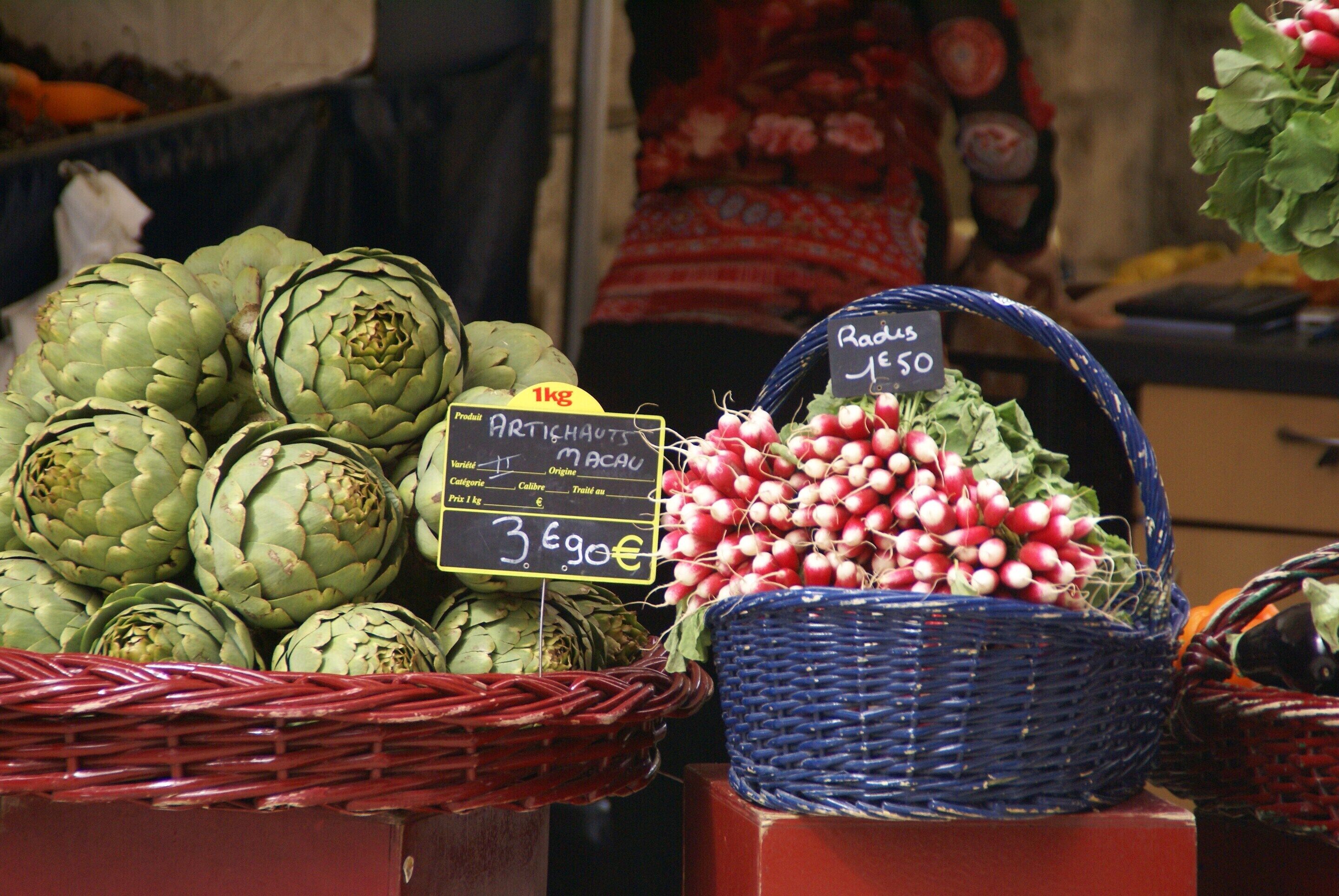 Un étal de marché où l’on trouve des paniers d’artichauts et de radis, avec des étiquettes indiquant les prix.