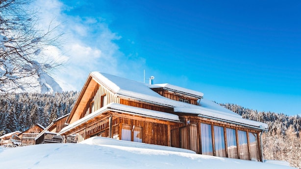 Un chalet en bois, doté d’un toit à forte pente, entouré d’arbres couverts de neige et surplombé par un ciel bleu dégagé.