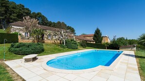 Ein Schwimmbecken mit einer Steindeck, umgeben von einer gepflegten Rasenfläche und einem Steinhaus.