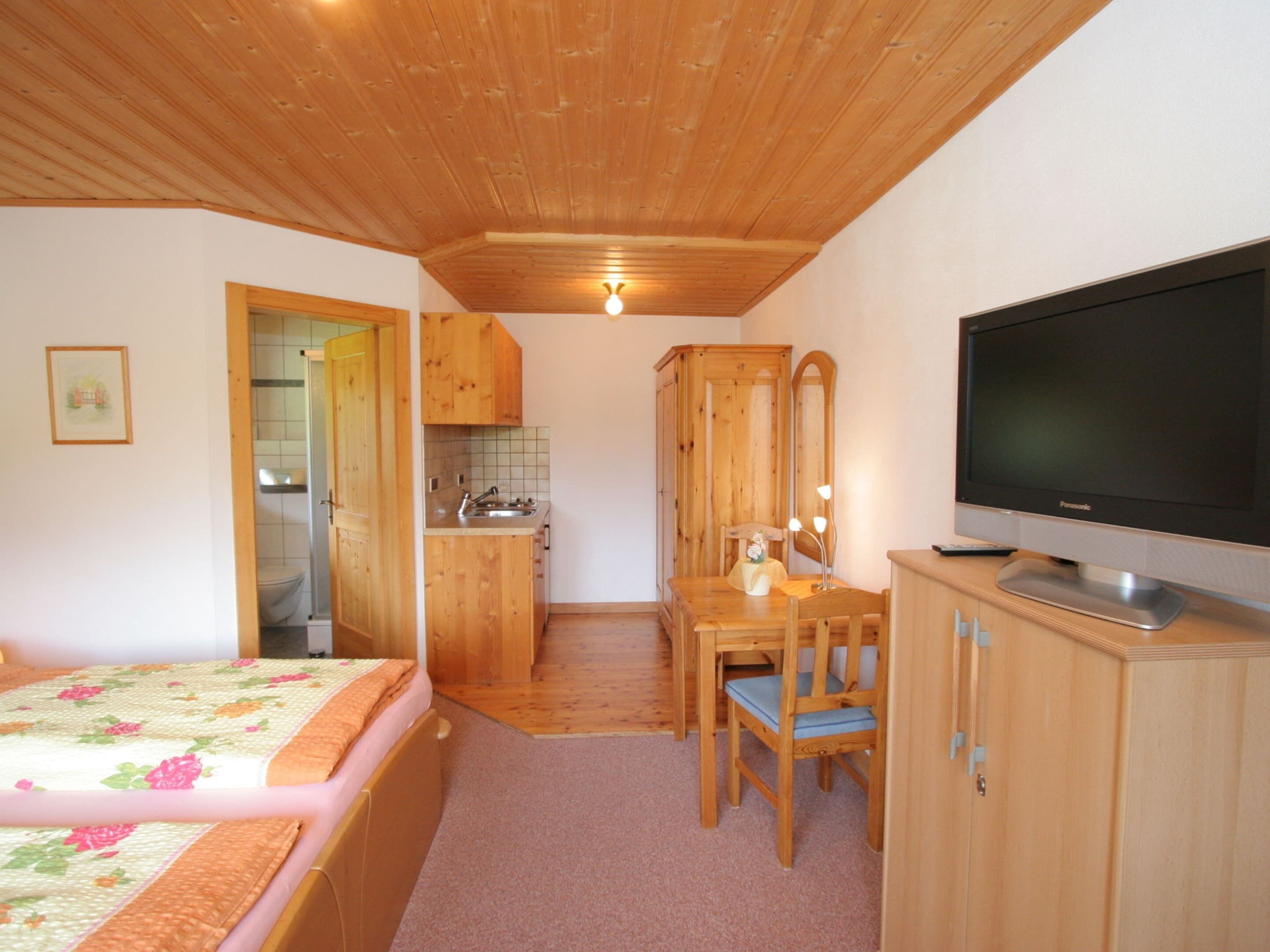 A compact room with a bed, a wooden dining table with chairs, a television, and a kitchenette with wooden cabinets.