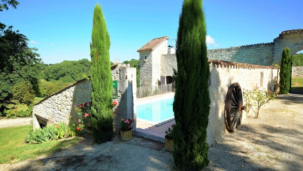 A stone building with a swimming pool, surrounded by greenery and a clear sky.