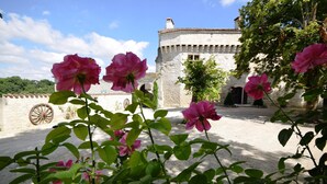 A stone building with a wheel in the foreground, surrounded by greenery and pink flowers.