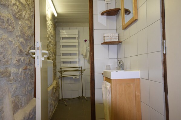 Ein schmaler Gang mit einer Steinsäule auf einer Seite, einer weiß gefliesten Wand auf der anderen, einem hölzernen Türrahmen und einem weißen Waschbecken mit Spiegel darüber.