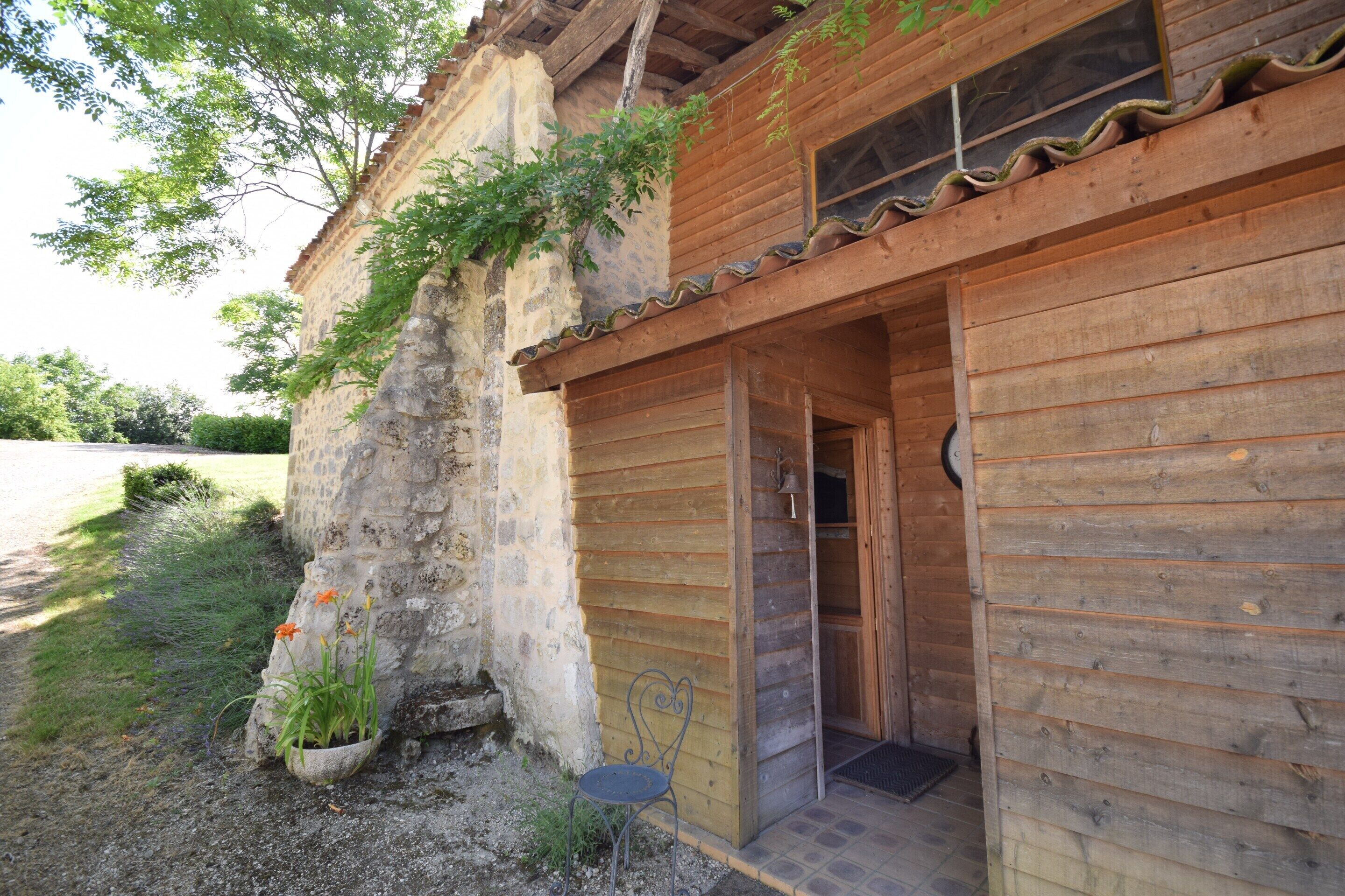 Un edificio rústico de madera con un muro de piedra, una puerta de madera y un pequeño jardín con un banco.