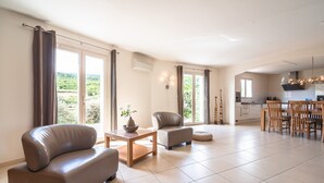 Una sala de estar moderna con sofá, sillón y mesa de comedor, una cocina al fondo y ventanas grandes con cortinas.