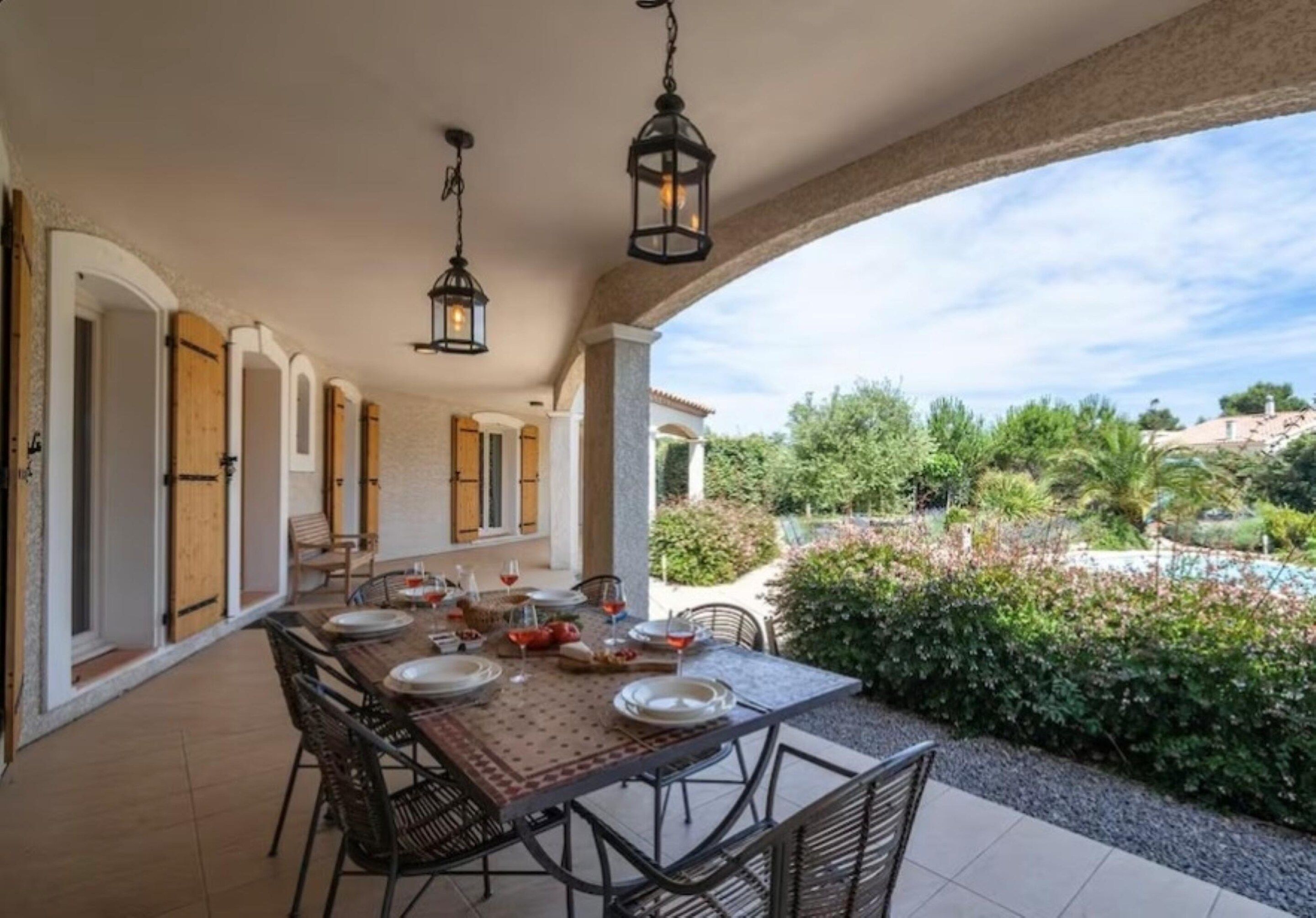 An outdoor dining area with a table set for four, surrounded by chairs, under a covered patio with hanging lanterns.