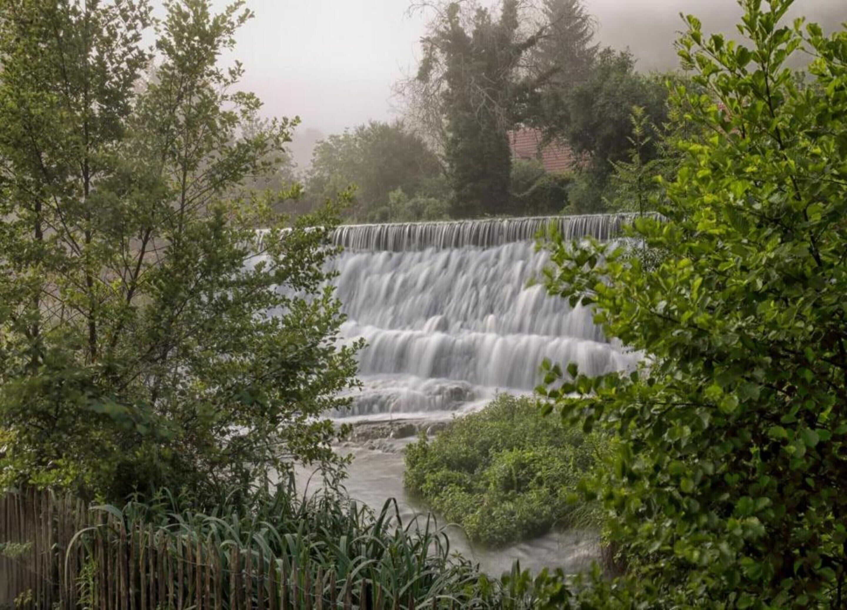 Uma cascata despencando sobre um paredão rochoso, cercada por vegetação exuberante e árvores.