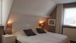 A bedroom with a sloped ceiling, a bed with two pillows, bedside lamps, a framed picture on the wall, and a window with blinds. - Groet Villa Stay With Garden Views (Groet)