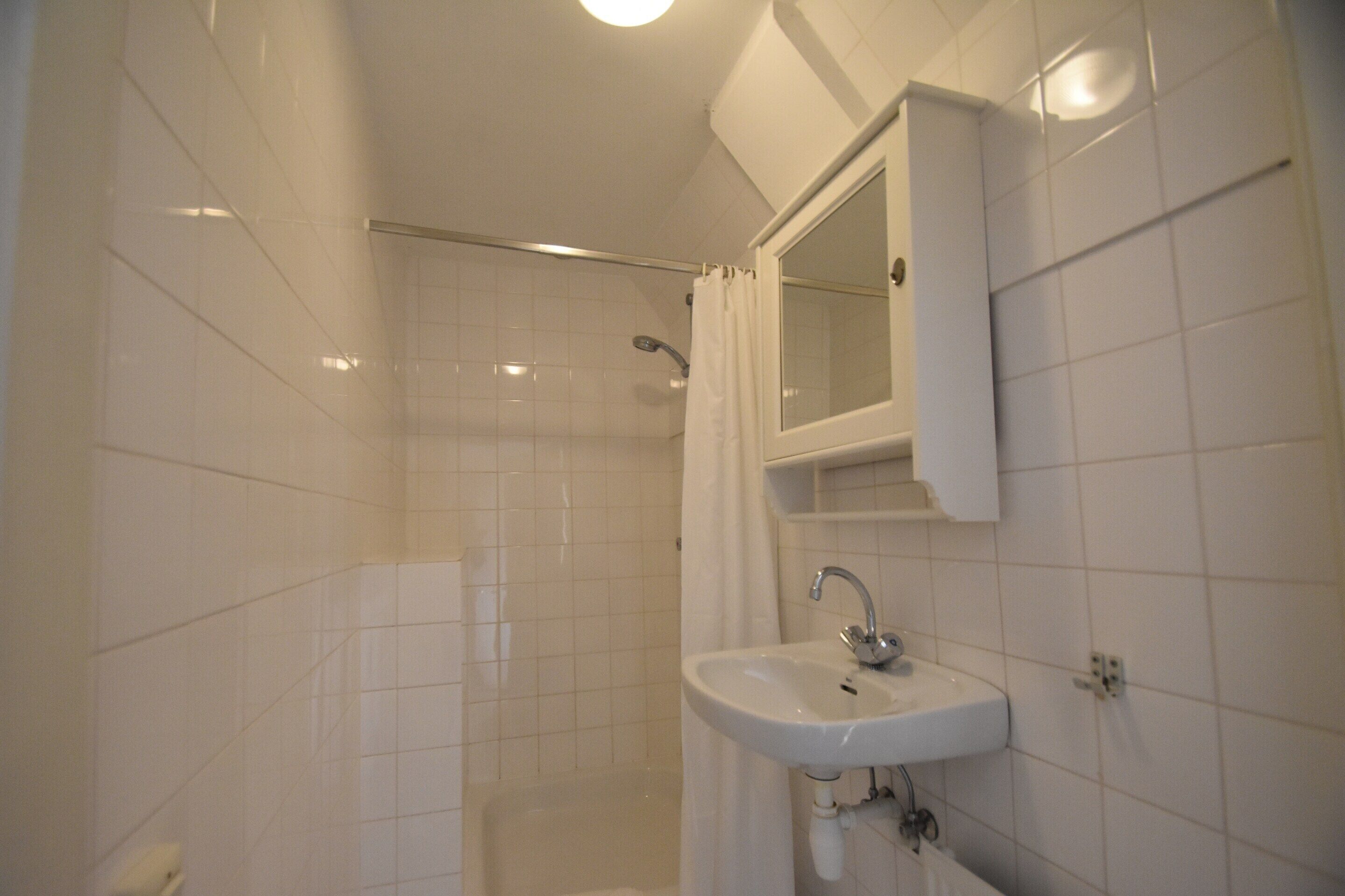 A bathroom with white tiles, a sink, a mirror, and a shower area.