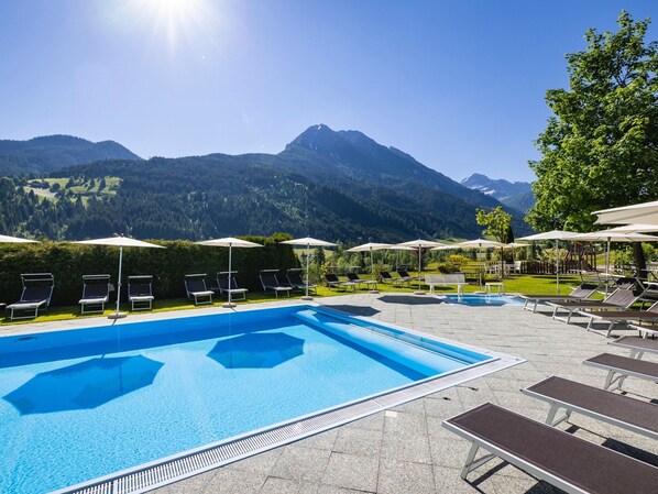 A swimming pool with lounge chairs and umbrellas, surrounded by mountains and greenery. - Apartment in Kleinarl Near Ski Slopes (Kleinarl)