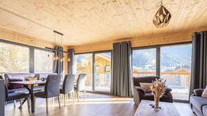 Amplia sala de estar con techo de madera, ventanas grandes que ofrecen vista a la montaña, comedor con mesa y sillas, y un sofá con un jarrón de flores.