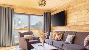 Una sala de estar moderna con techo de madera, un televisor de pantalla plana grande montado en la pared, un juego de sofá, una mesa de centro y vistas al exterior a través de amplios ventanales.