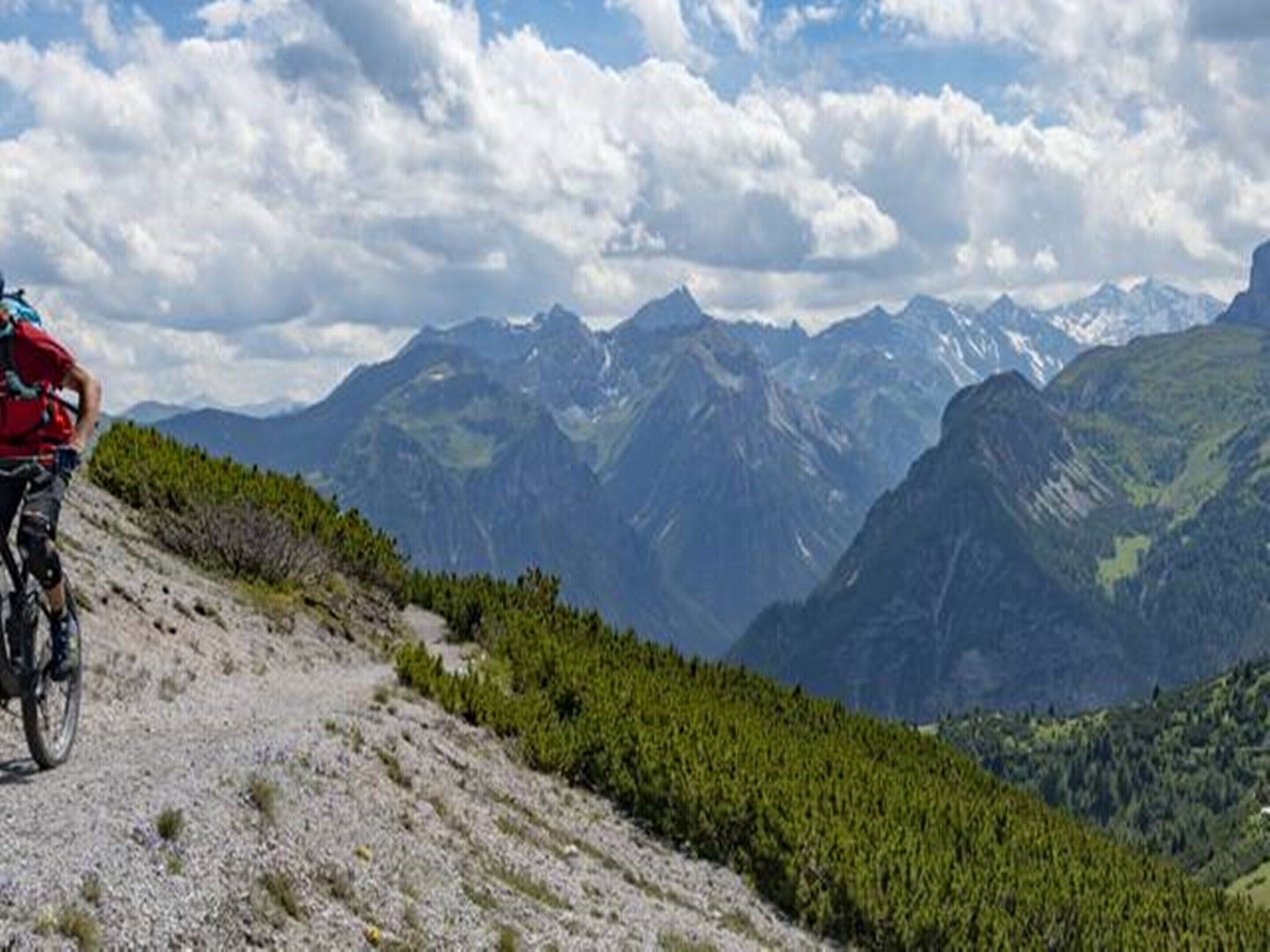 Ein Mountainbiker auf einem Schotterweg mit Blick auf schneebedeckte Berge und grüne, bewaldete Hänge.