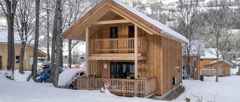 A two-story wooden cabin with a balcony, surrounded by snow-covered trees and a mountainous backdrop.