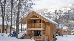 A two-story wooden cabin with a balcony, surrounded by snow-covered trees and a mountainous backdrop.