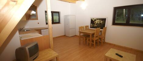 A small, open-plan room with a wooden ceiling, a kitchenette, a dining area with chairs, a television on a wooden table, and a refrigerator.
