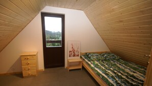 Quarto com teto inclinado, cama com colcha estampada, cómoda de madeira pequena e porta que dá para uma varanda.