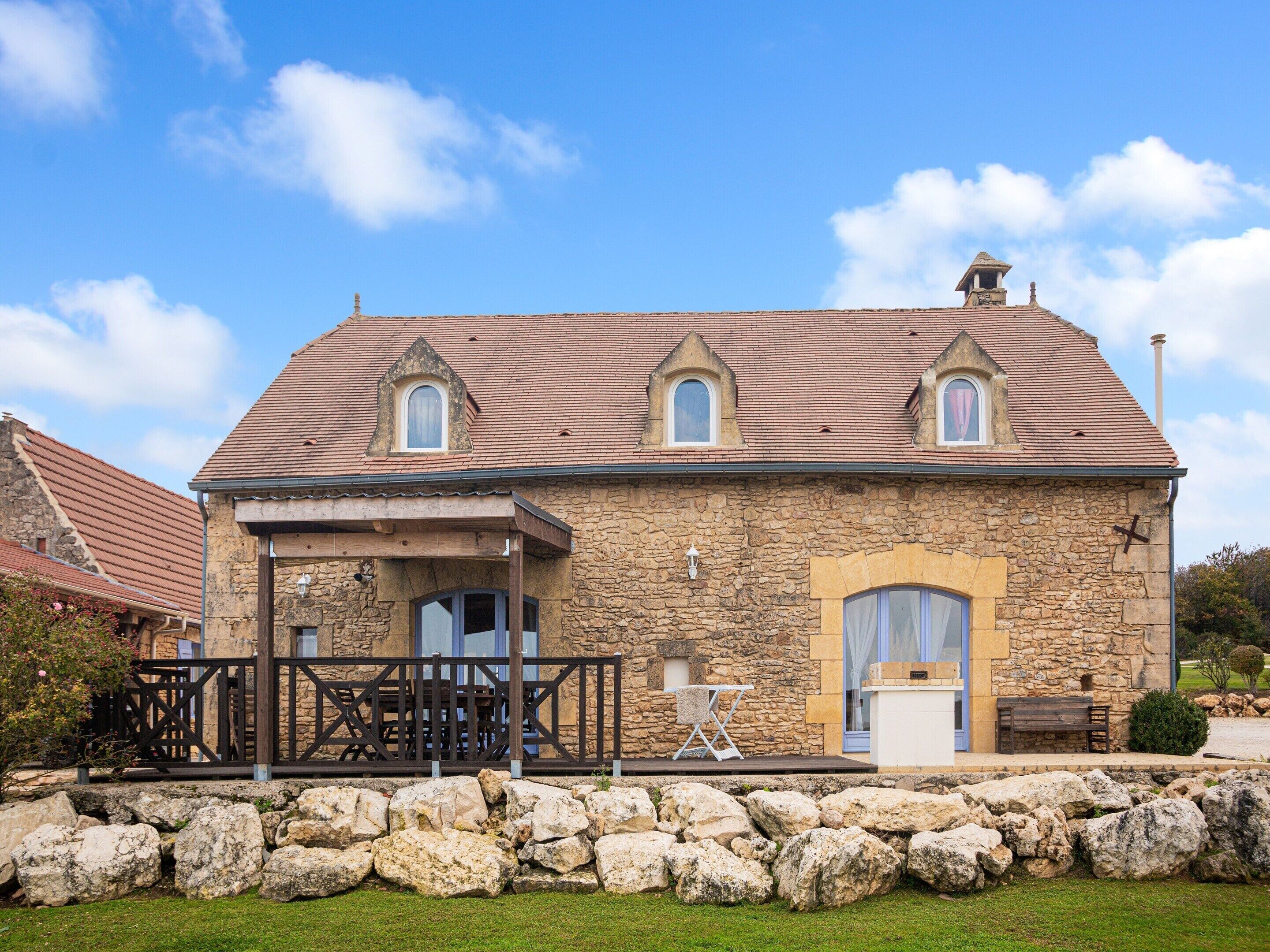 A stone building with a tiled roof, a covered patio with outdoor furniture, and a stone wall in front.