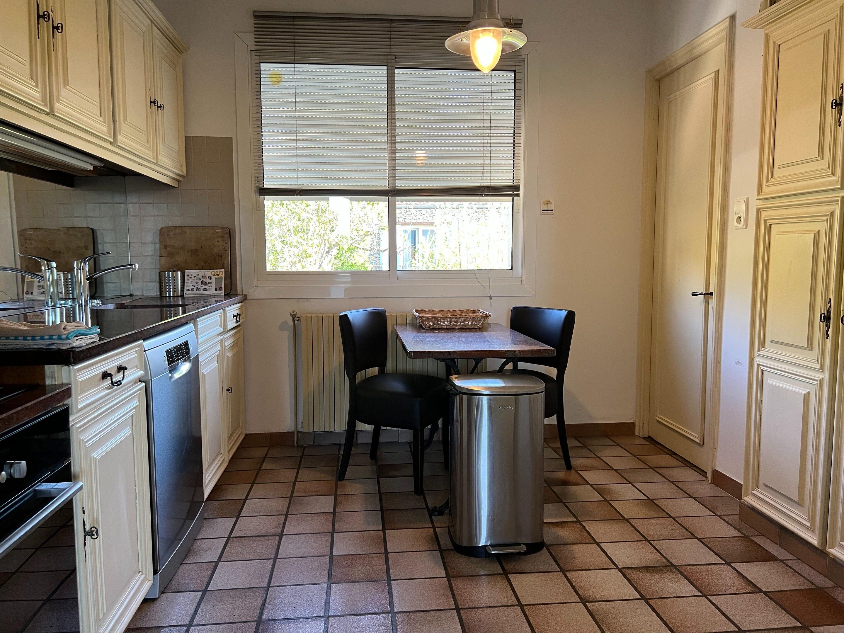 A kitchen with a dining table, chairs, a trash bin, and cabinets.