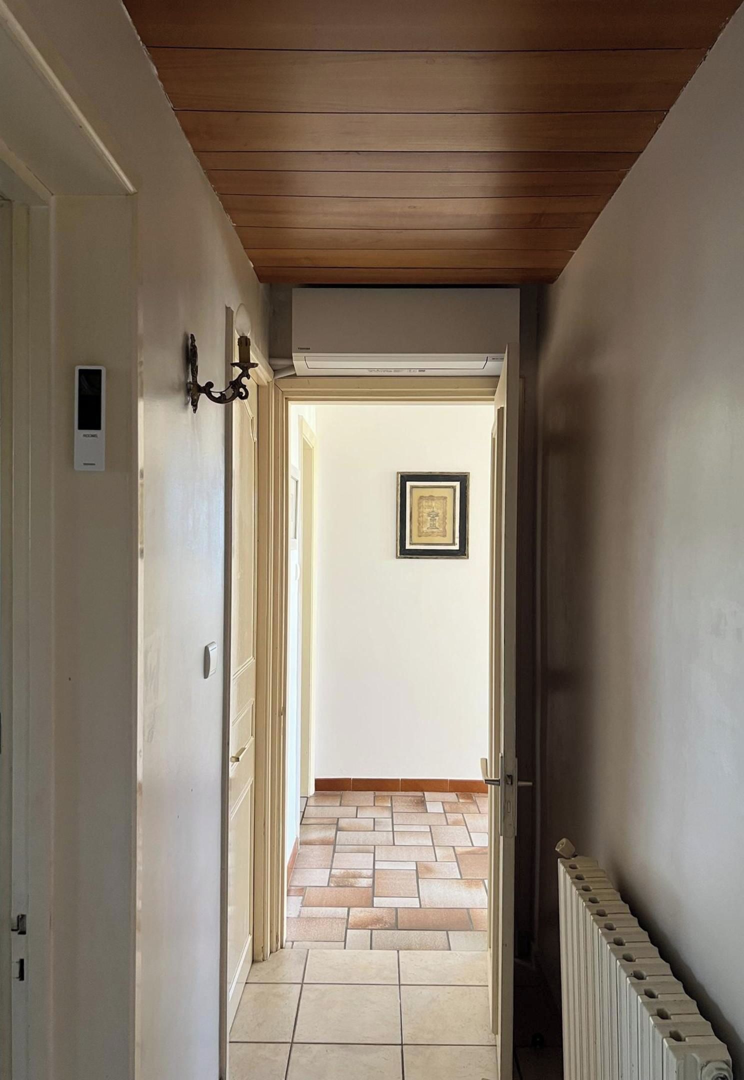 A hallway with a wooden ceiling, a radiator, and a tiled floor leading to a room with a door.