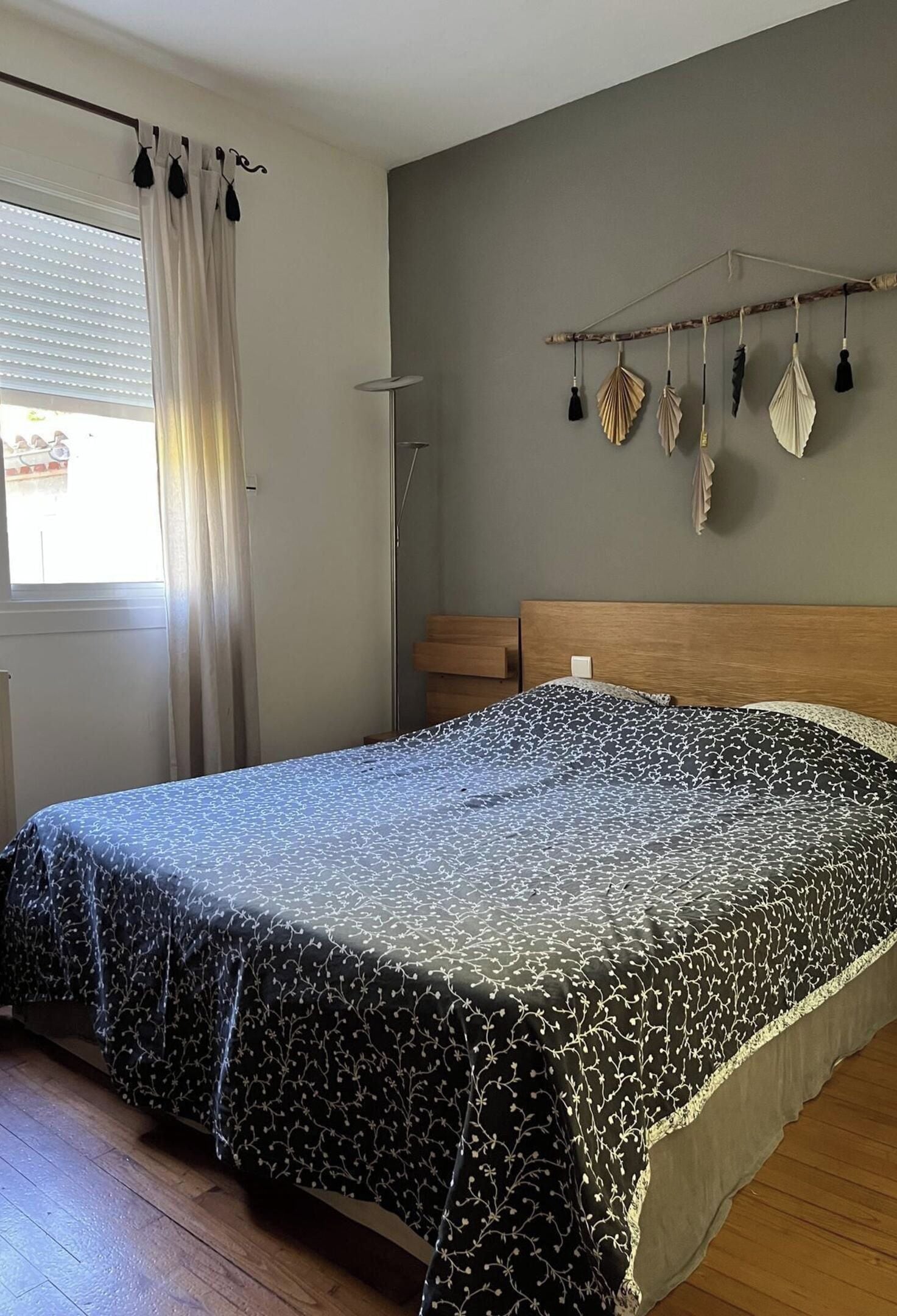 A bedroom with a bed, a window with curtains, a bedside table, and a wall-mounted light fixture.