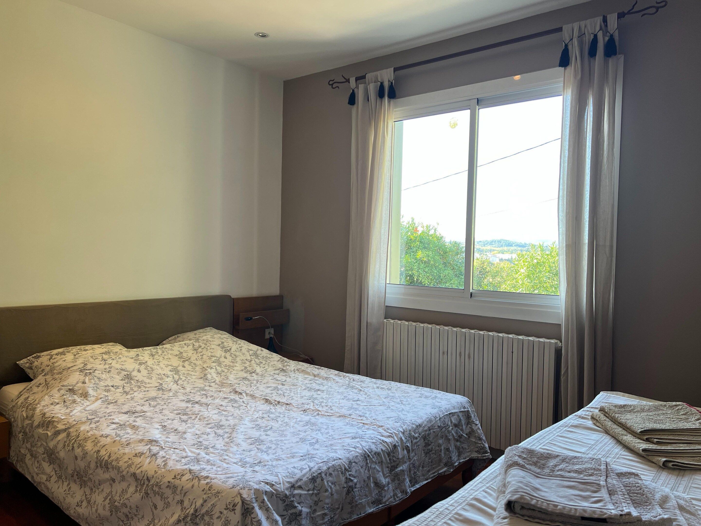 A bedroom with a bed, a nightstand, a window with curtains, and a radiator.