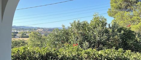 Vista a través de un ventanal arqueado sobre un jardín exuberante y un pueblo lejano.