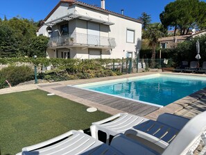 Una piscina con sillones de descanso, un jardín cercado y un edificio de dos pisos con balcón.