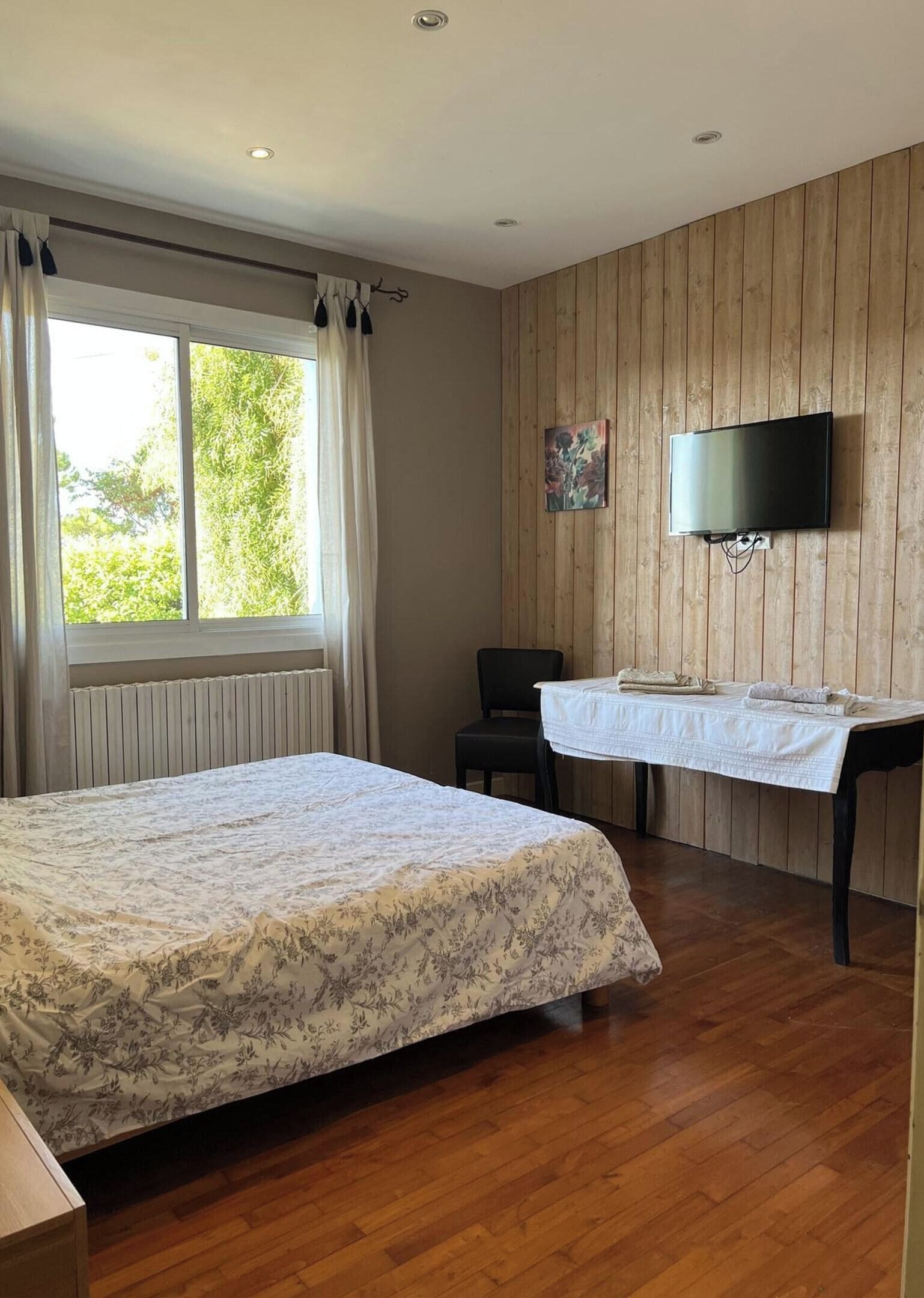 A bedroom with a bed, a desk, a chair, a television, and a window with curtains.