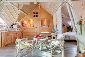 A rustic wooden cabin with a loft bedroom, a dining area with wooden chairs, and a kitchen area with a brick wall. - Mountain Stay Near Canal (Niderviller)