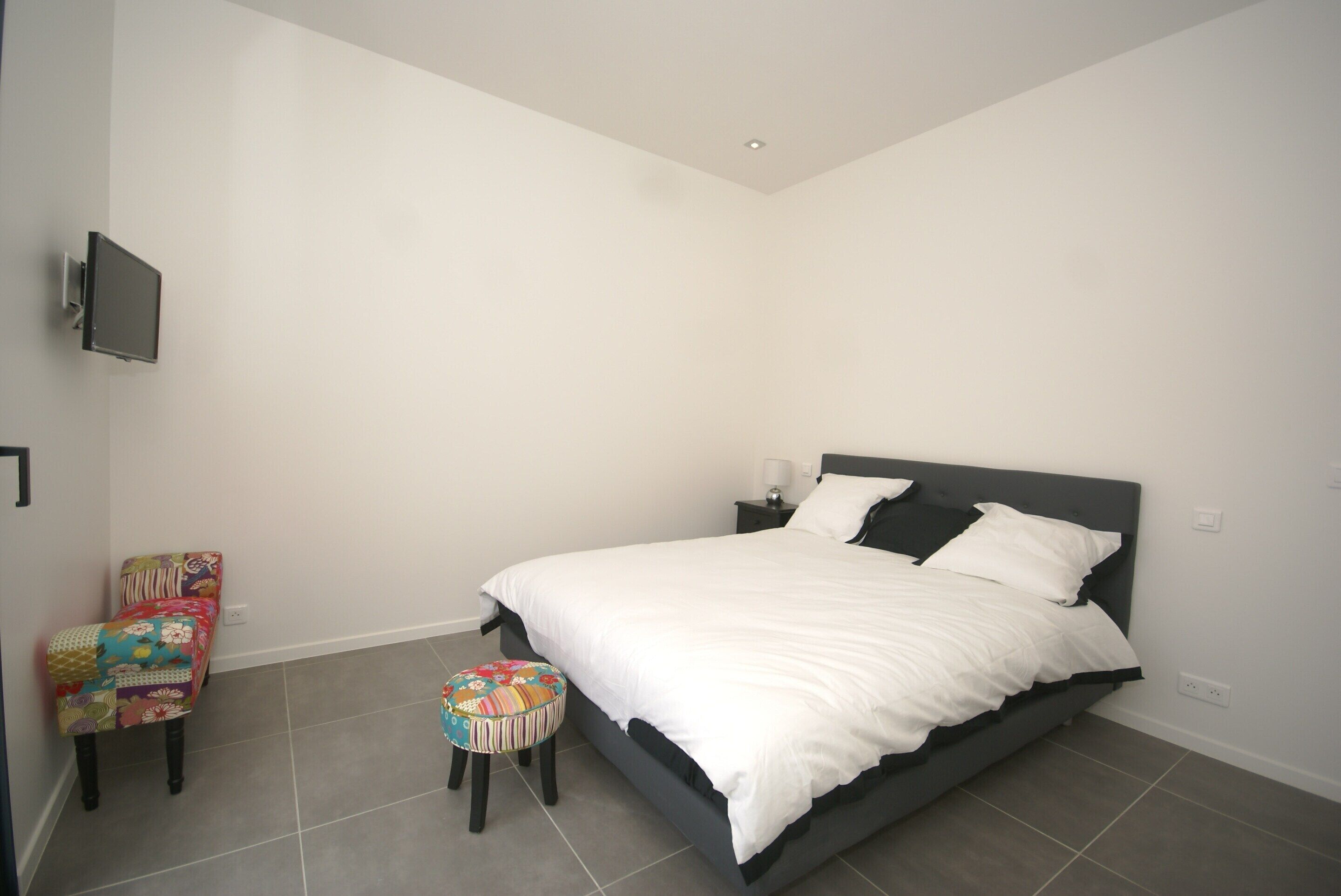 A bedroom with a bed, a chair, a stool, a television, and a nightstand.