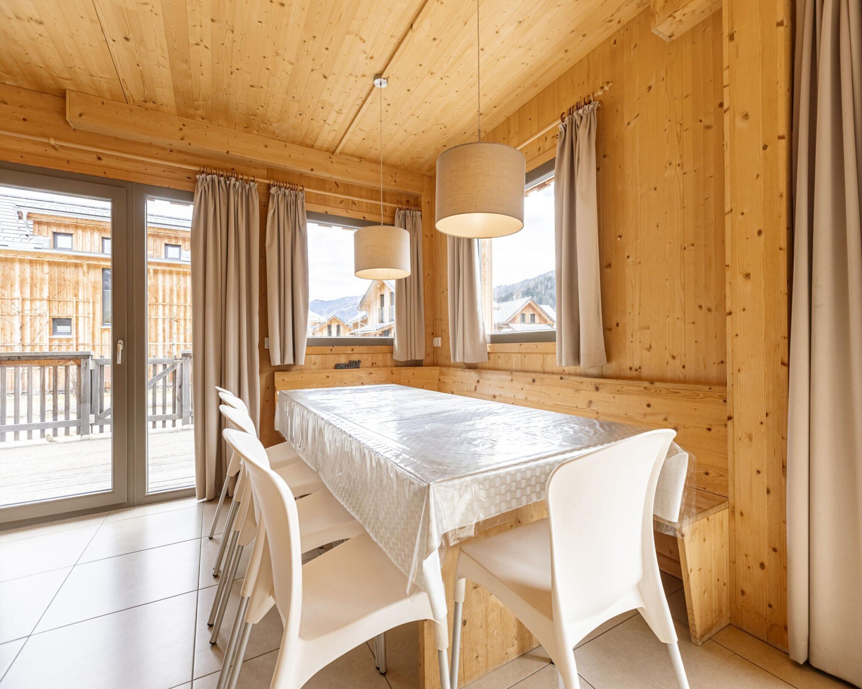 A wooden cabin with a dining table and chairs, large windows, and a view of buildings outside.