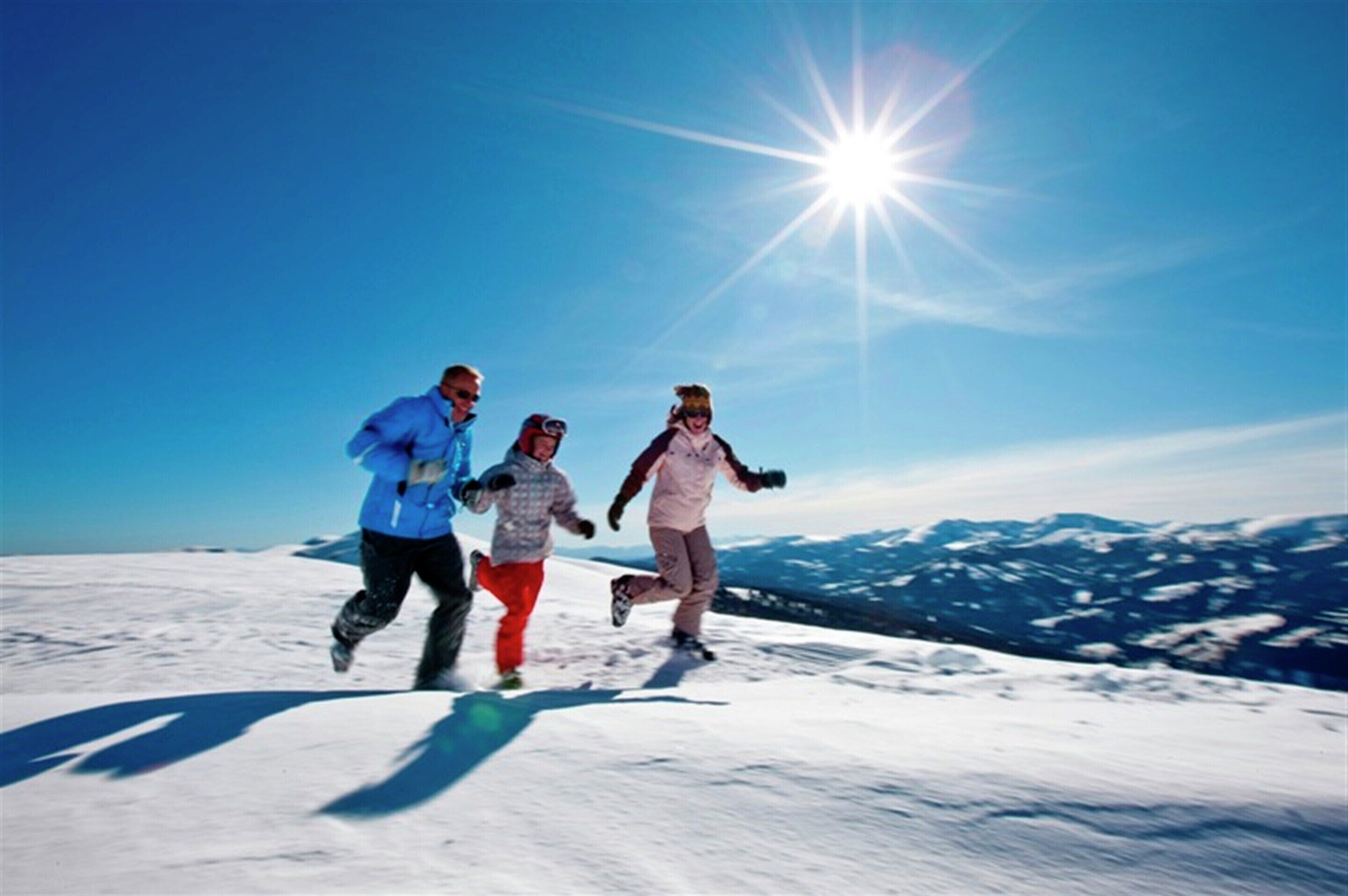 Drei Personen laufen auf einem schneebedeckten Berg, im Hintergrund ein klarer Himmel und Sonne.