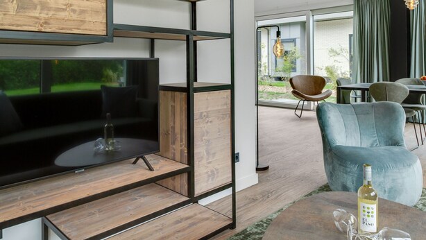 A modern living room with a large flat-screen TV, wooden shelving, a blue armchair, and a dining area with a table and chairs. - Bungalow in Texel Near National Park Dunes (De Koog)