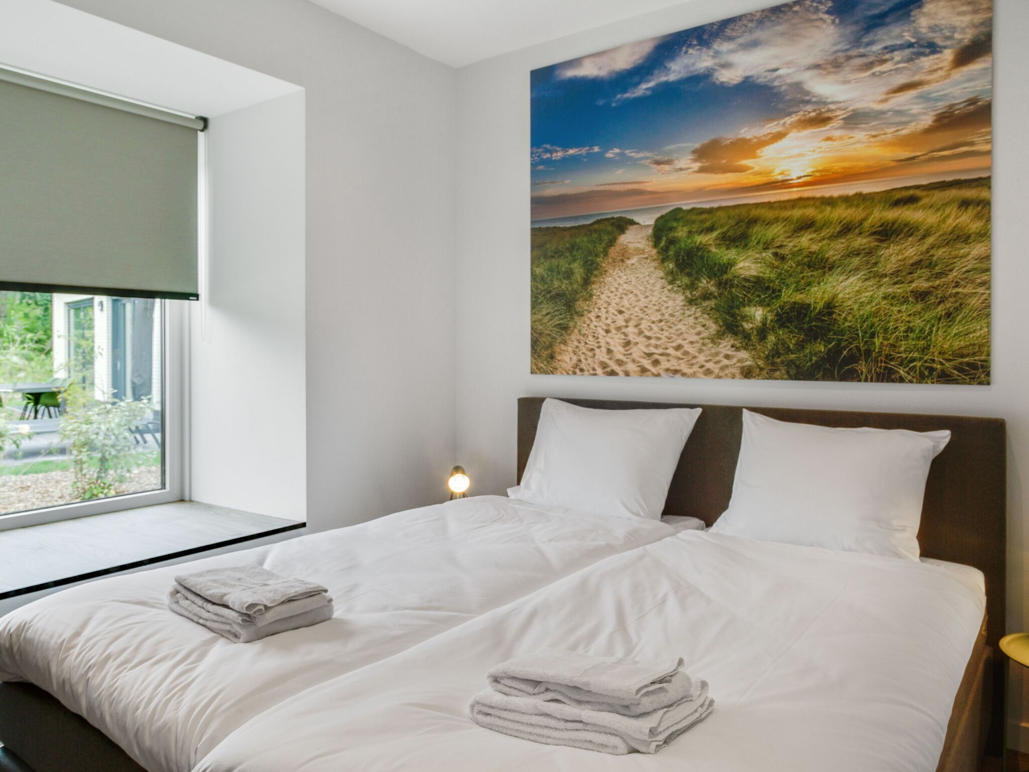 A bedroom with a large bed, a window with a view of the outdoors, and a painting of a beach scene on the wall.