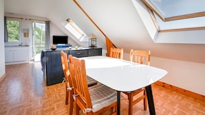 A dining area with a white table and wooden chairs, a kitchen in the background, and a sloped ceiling with skylights.