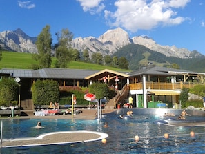 Una piscina con gente, un edificio con balcón y montañas al fondo.