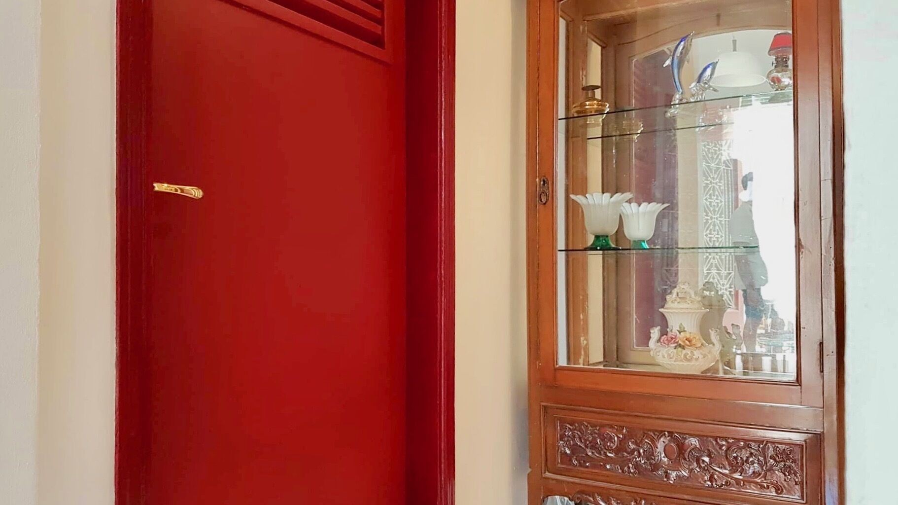A red door with a brass handle, a glass-fronted cabinet with decorative items, and a reflection of a person in the glass.