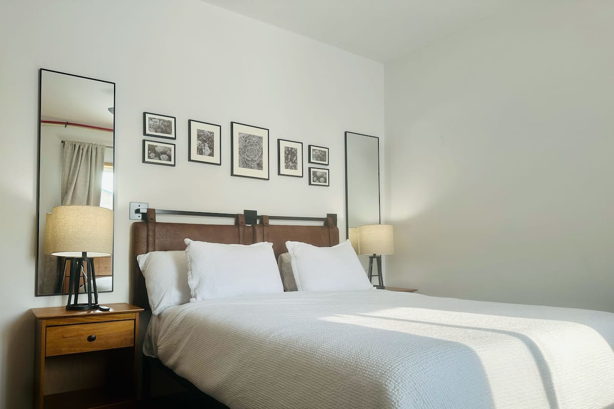 A neatly made bed with a white comforter and pillows, a wooden nightstand with a lamp, and a wall with framed pictures.