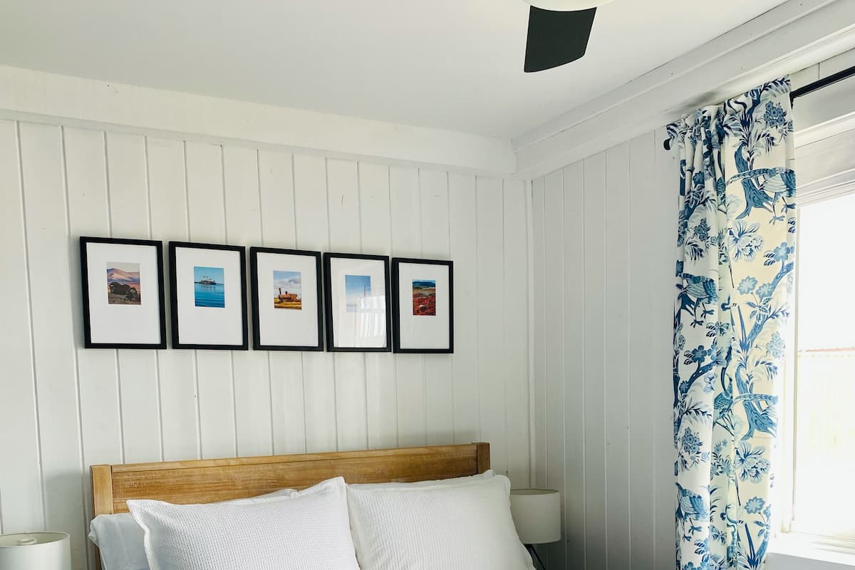 A bedroom with a wooden headboard, white bedding, a nightstand with a lamp, and framed pictures on the wall.