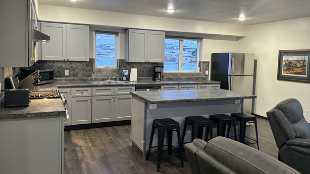 A modern kitchen with white cabinets, a granite countertop, and a central island with bar stools. - Noxapaga Suites (Nome)