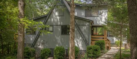 A rustic cabin with a steeply pitched roof, surrounded by dense forest. A wooden deck leads to the entrance, and a small porch is visible on the right side.