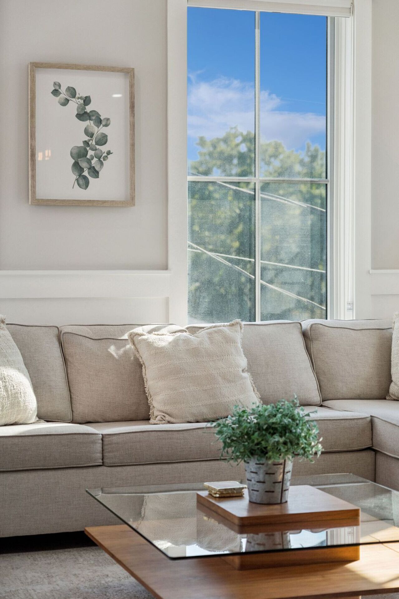 Un salon avec un canapé d’angle beige, une table basse en verre, une estampe botanique encadrée accrochée au mur et une vue sur les arbres par une grande fenêtre.