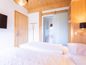 Quarto com teto de madeira, cama com roupa de cama branca, porta de madeira e televisão presa à parede.