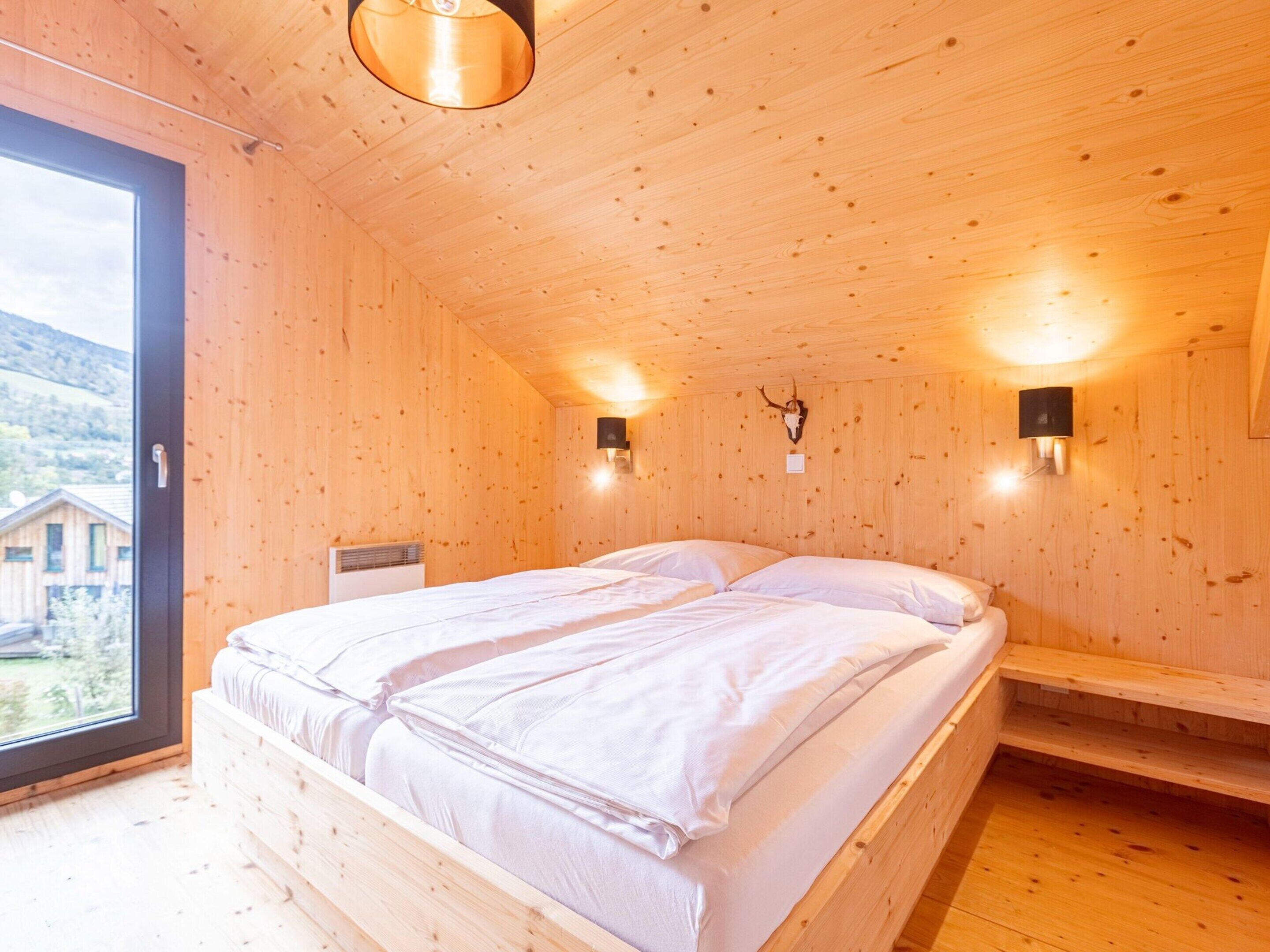 A wooden cabin bedroom with a bed, two wall-mounted lights, and a view of the outside.