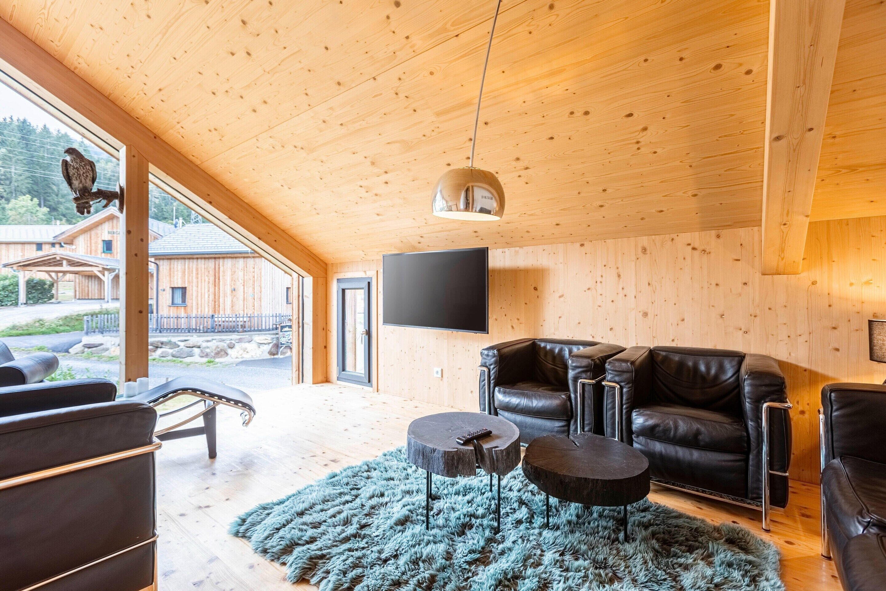 Un salon moderne avec un plafond en bois, des canapés en cuir, un téléviseur à écran plat et une grande fenêtre donnant sur un chalet en bois et un espace extérieur.