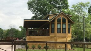 A small wooden cabin with a balcony, surrounded by a fence and trees.