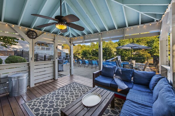 A covered patio area with a ceiling fan, string lights, a seating area with a sofa and chairs, and a view of a backyard with a pool and lounge chairs.