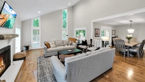 A spacious living room with a fireplace, a large grey sofa, a coffee table, and a dining area with a chandelier. - Lake Norman Luxury - Large Saltwater Pool, Dock & Boathouse - Sleeps 18 (Denver)